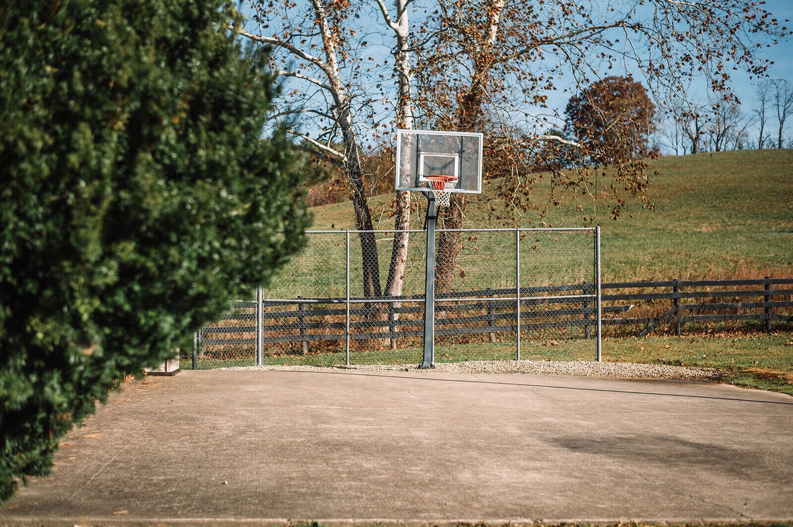 An outdoor basketball court with a chain-link fence, trees, and a grassy hill in the background.