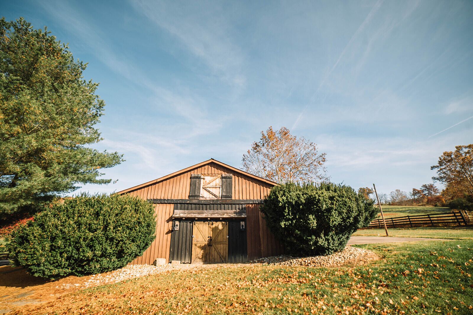 Une grange en bois avec un toit à pignon, un grand arbre sur la gauche et un champ clôturé en arrière-plan.