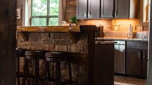 A rustic kitchen with wooden beams, a stone island, bar stools, and a view of the outdoors.