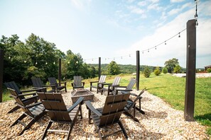 Un espace extérieur aménagé avec des chaises Adirondack et un foyer, entouré d’une terrasse en gravier et illuminé de guirlandes lumineuses.