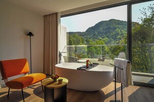 Un baño moderno con una bañera independiente, una silla, una mesita con fruta y vistas a una ladera boscosa.