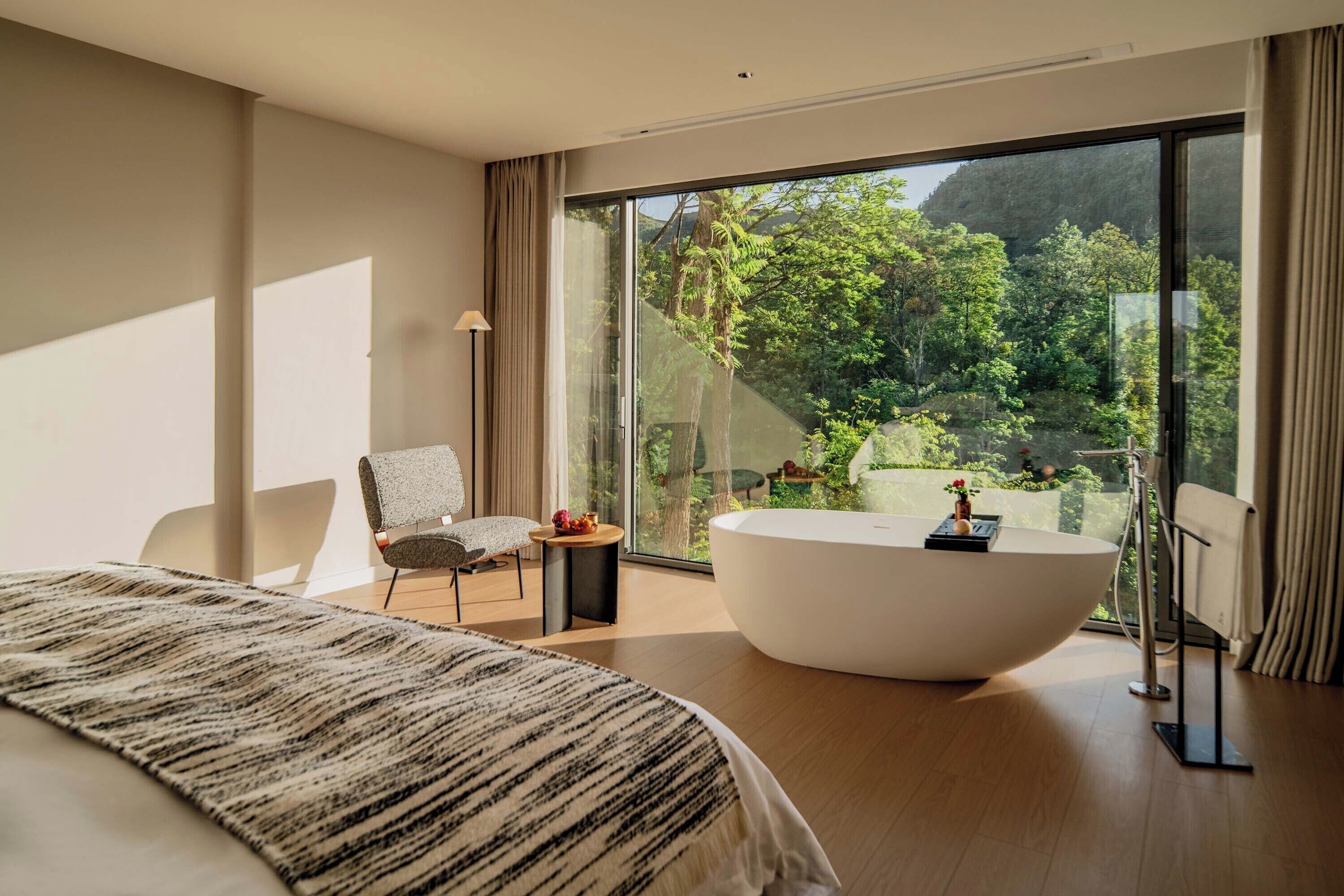 Un dormitorio moderno con una cama grande, una zona de estar con una silla, una bañera y vistas a un bosque a través de ventanas amplias.