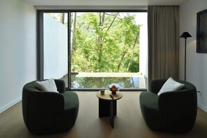 Una habitación moderna con dos sillones verdes, una mesita redonda con fruta y un ventanal con vistas a árboles.