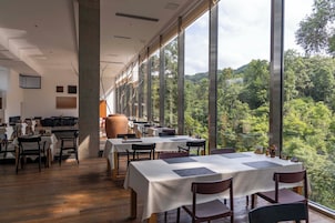 Restaurante com mesas e cadeiras dispostas no interior, grandes janelas oferecendo vista para um morro arborizado.