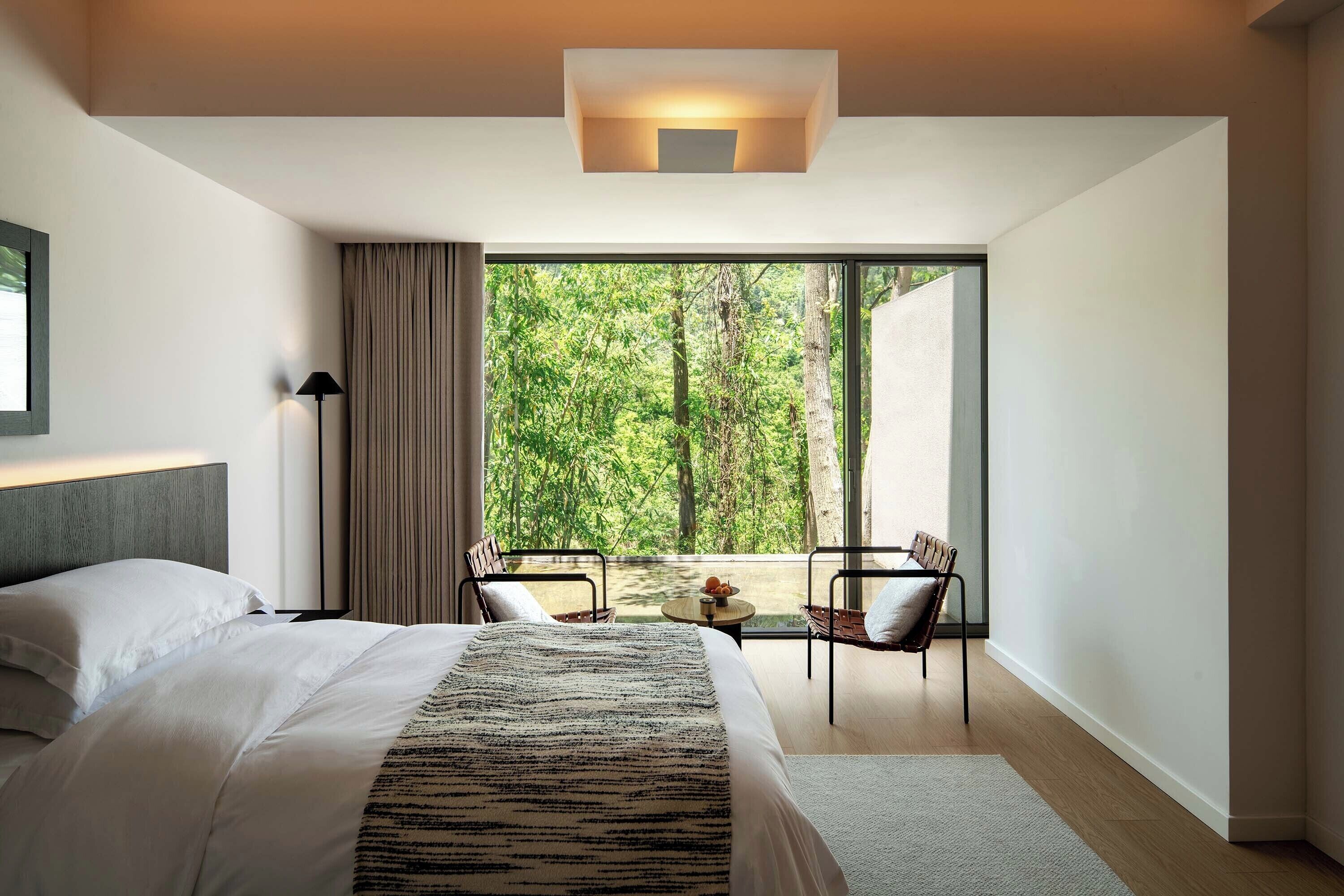 Un dormitorio moderno con una cama grande, vistas a zonas verdes a través de un ventanal corredera y una pequeña mesa con sillas.