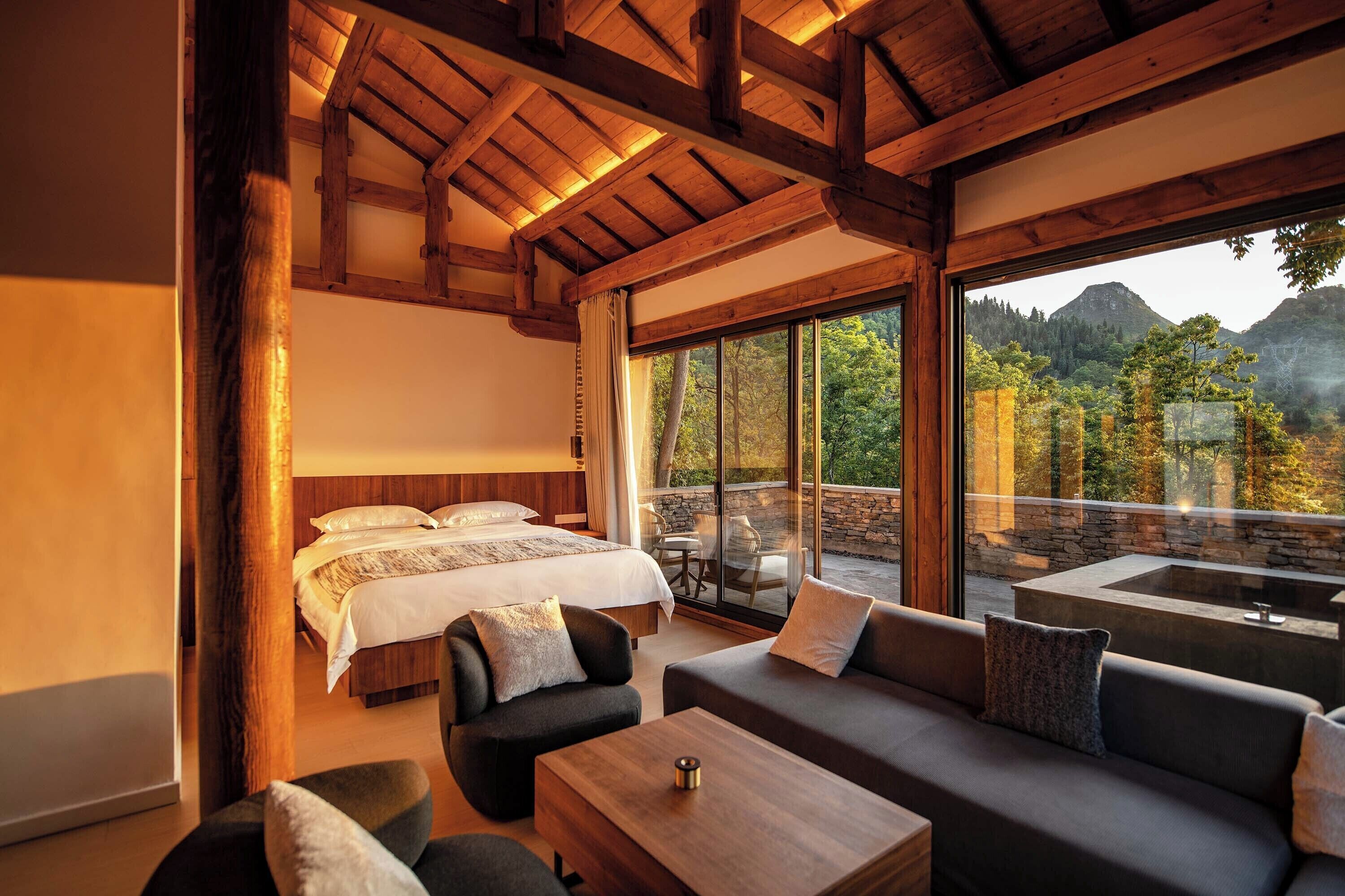 A bedroom with a large bed, a sofa, and a coffee table. There is a view of a mountainous landscape through large windows.