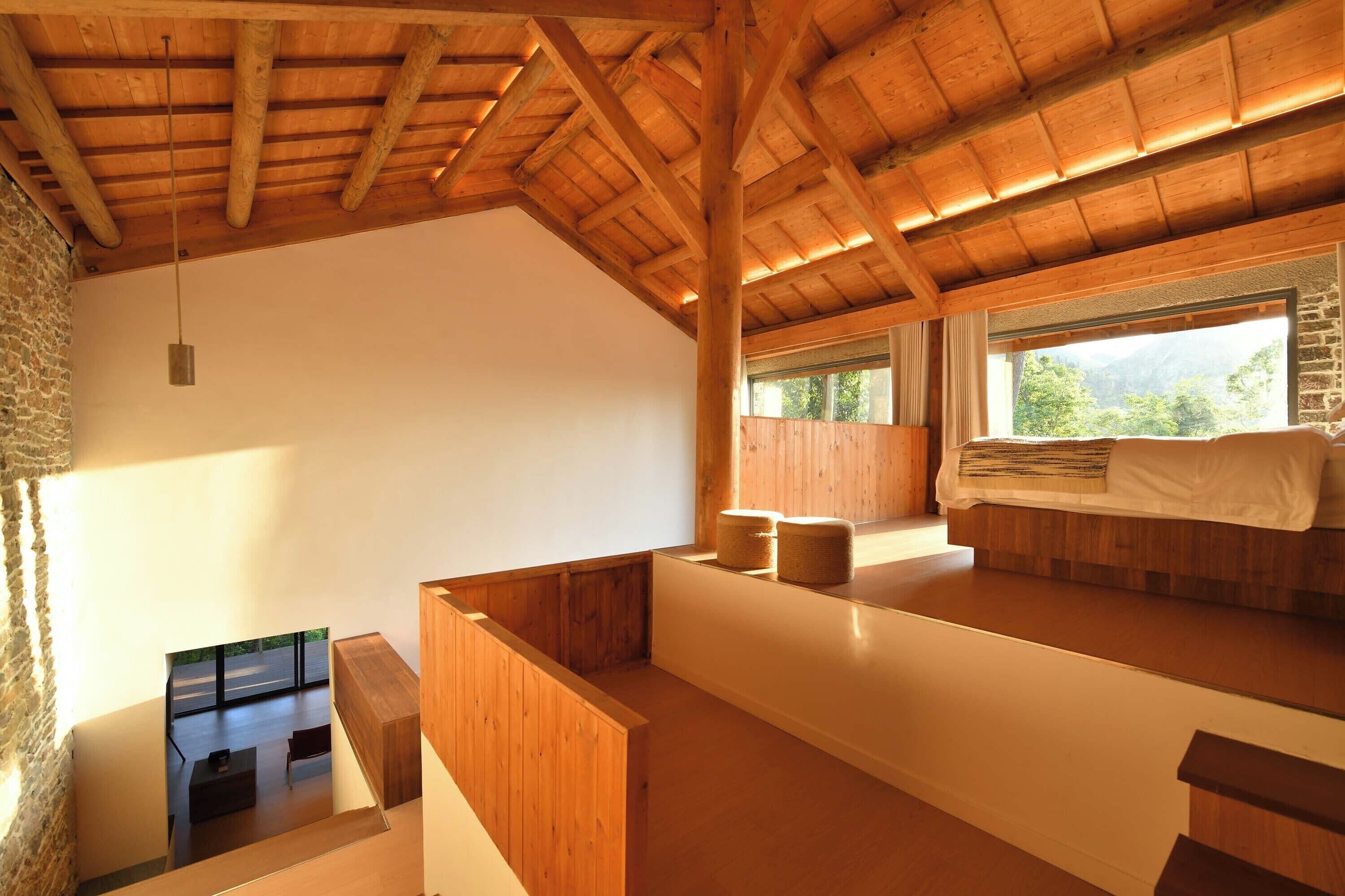 Una habitación con techo de madera inclinado, una cama con cubrecama blanco, un ventanal con vistas a zonas verdes y una pequeña mesa con una silla.