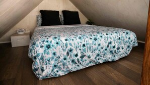 A bedroom with a sloped ceiling, a bed with a floral bedspread, two black pillows, a bedside table with a lamp, and a small plant.