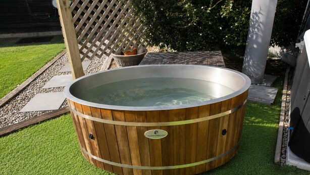 A wooden hot tub with a metal rim, surrounded by artificial grass, a wooden fence, and a potted plant.