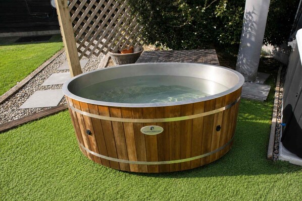 Una bañera de madera con borde metálico, rodeada de césped artificial, una cerca de madera y una planta en maceta.