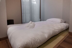 A neatly made bed with white linens and two rolled towels, a window with blinds, and a wooden floor. - Kikou (Kyoto)