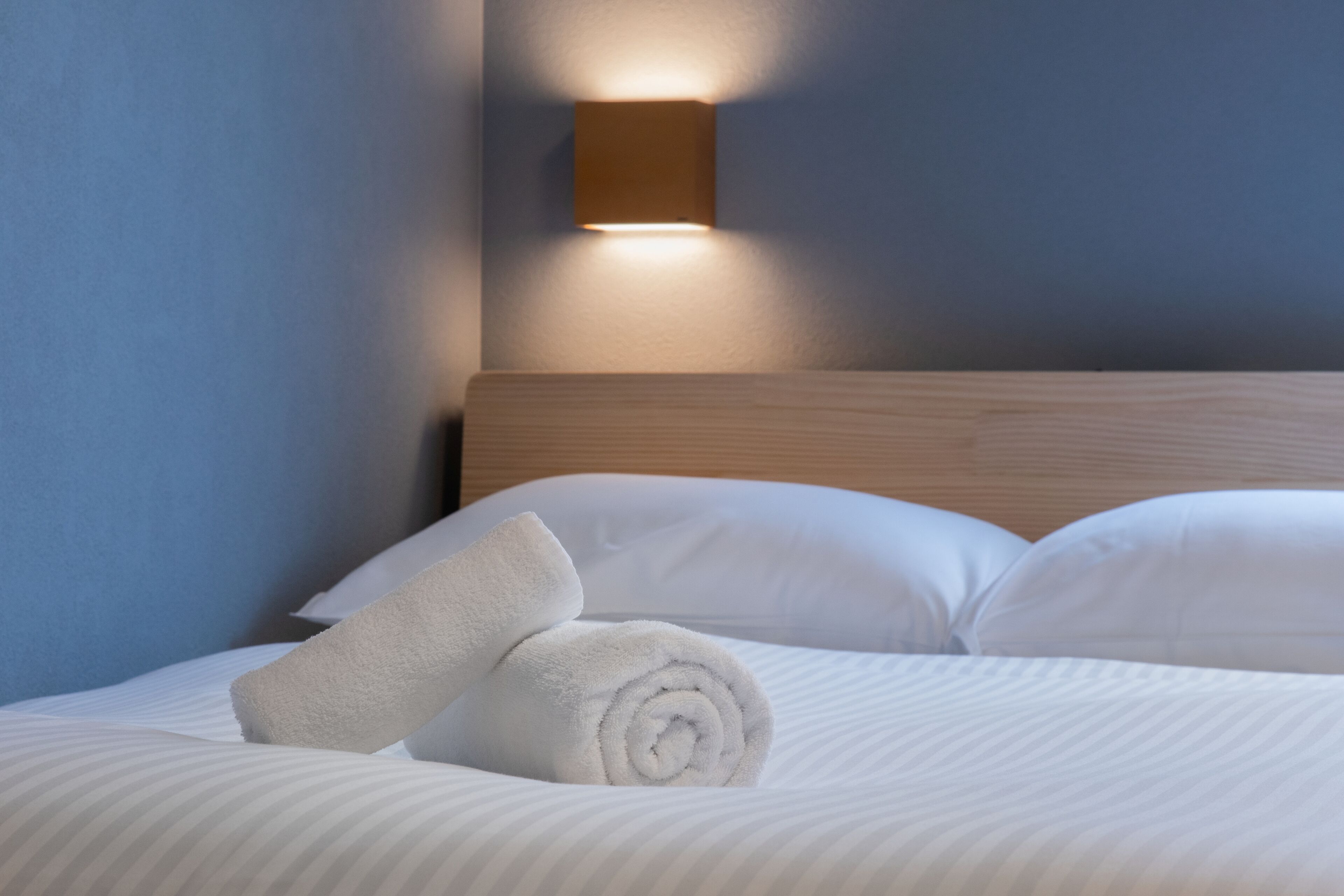 A neatly made bed with white linens and pillows, a rolled white towel, and a wall-mounted light fixture.