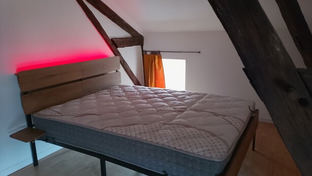 Un lit simple dans une chambre dont le plafond est en pente et où les poutres en bois sont apparentes.