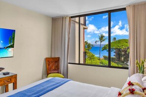 A hotel room with a bed, a desk, a chair, a window with a view of the ocean and greenery, and a painting on the wall. - Large 1BR condo, partial ocean view, pool, beach access - Kaanapali Shores 414 (Lahaina)