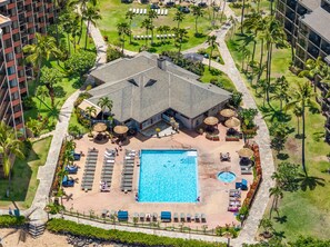 An aerial view of a hotel with a central swimming pool surrounded by lounge chairs and umbrellas, with palm trees and a lawn in the background. - Large 1BR condo, partial ocean view, pool, beach access - Kaanapali Shores 414 (Lahaina)