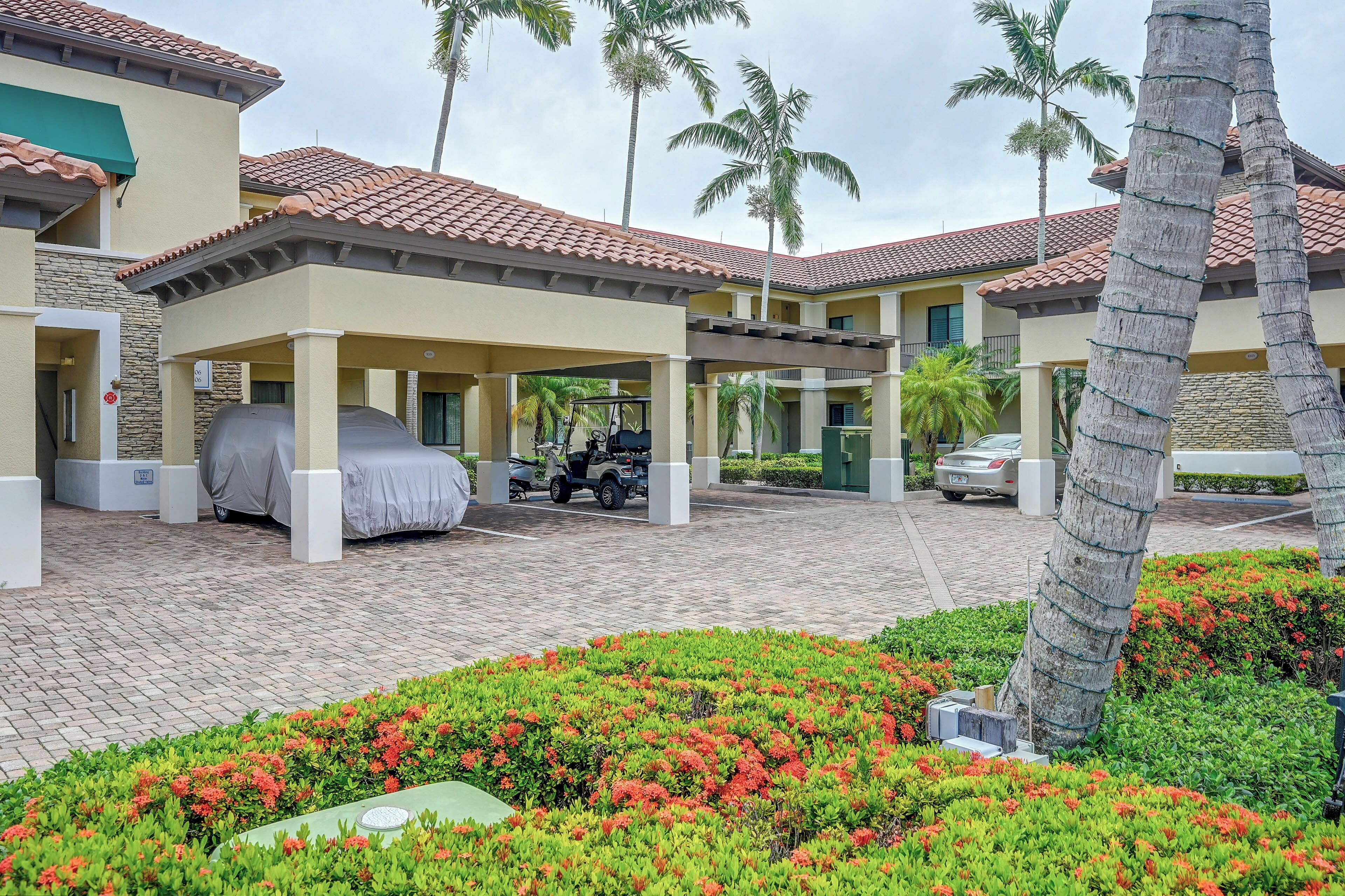 A covered outdoor area with a car and a covered vehicle, palm trees, and a building with a tiled roof.