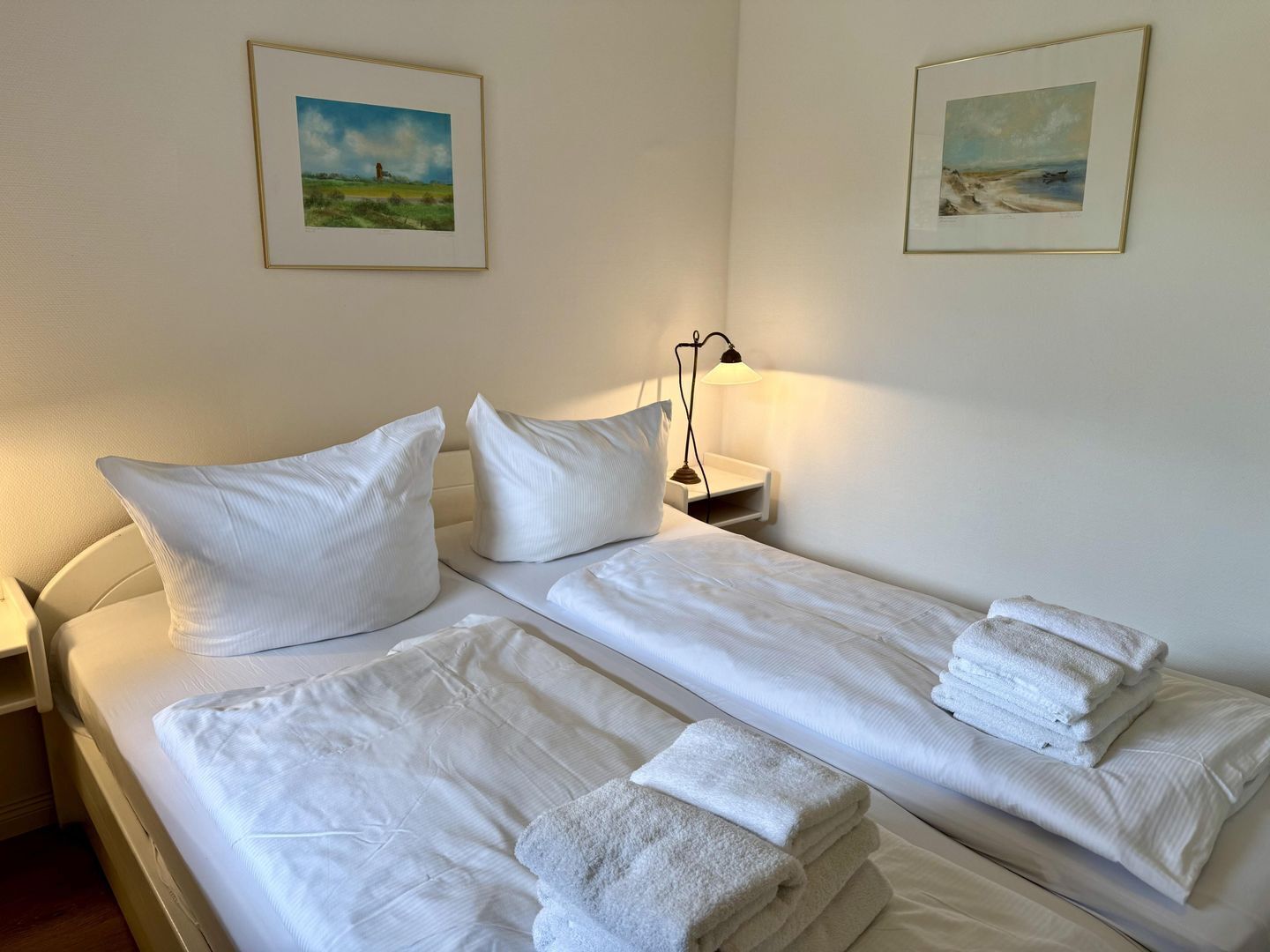 En dobbeltseng med hvide sengetøj og puder, en sengetårnslampe og to indrammede landskabsmalerier på væggen.
