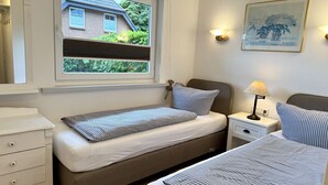 A bedroom with two single beds, a bedside table with a lamp, a dresser, and a window with a view of a house and greenery. - Reiherweg, Apartment 1 - Beautiful 2-room vacation apartment with a terrace, quiet and (Sylt)