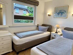 A bedroom with two single beds, a bedside table with a lamp, a dresser, and a window with a view of a house and greenery. - Reiherweg, Apartment 1 - Beautiful 2-room vacation apartment with a terrace, quiet and (Sylt)