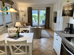 A compact kitchen with a dining area, a sofa, and a view through a glass door to an outdoor garden. - Reiherweg, Apartment 1 - Beautiful 2-room vacation apartment with a terrace, quiet and (Sylt)