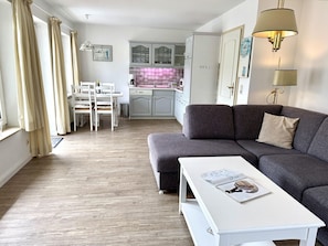 A modern living room with a grey sofa, a white coffee table with a magazine, a dining area with a white table and chairs, and a kitchen with light-colored cabinets. - Süderstraße, Apartment 10 – Landhaus - Cozy 2-room vacation rental for 4 (Sylt)