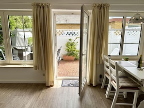 A room with a sliding glass door leading to a patio, a white dining table with chairs, and a small window with curtains. - Süderstraße, Apartment 10 – Landhaus - Cozy 2-room vacation rental for 4 (Sylt)