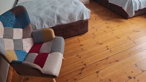 Et soveværelse med en stor seng, en mindre seng, en stol med pude i lapperi, et natbord med lampe og et maleri på væggen.