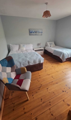 Ein Schlafzimmer mit einem großen Bett, einem kleineren Bett, einem Sessel mit Patchwork-Kissen, einem Nachttisch mit Lampe und einem Bild an der Wand.