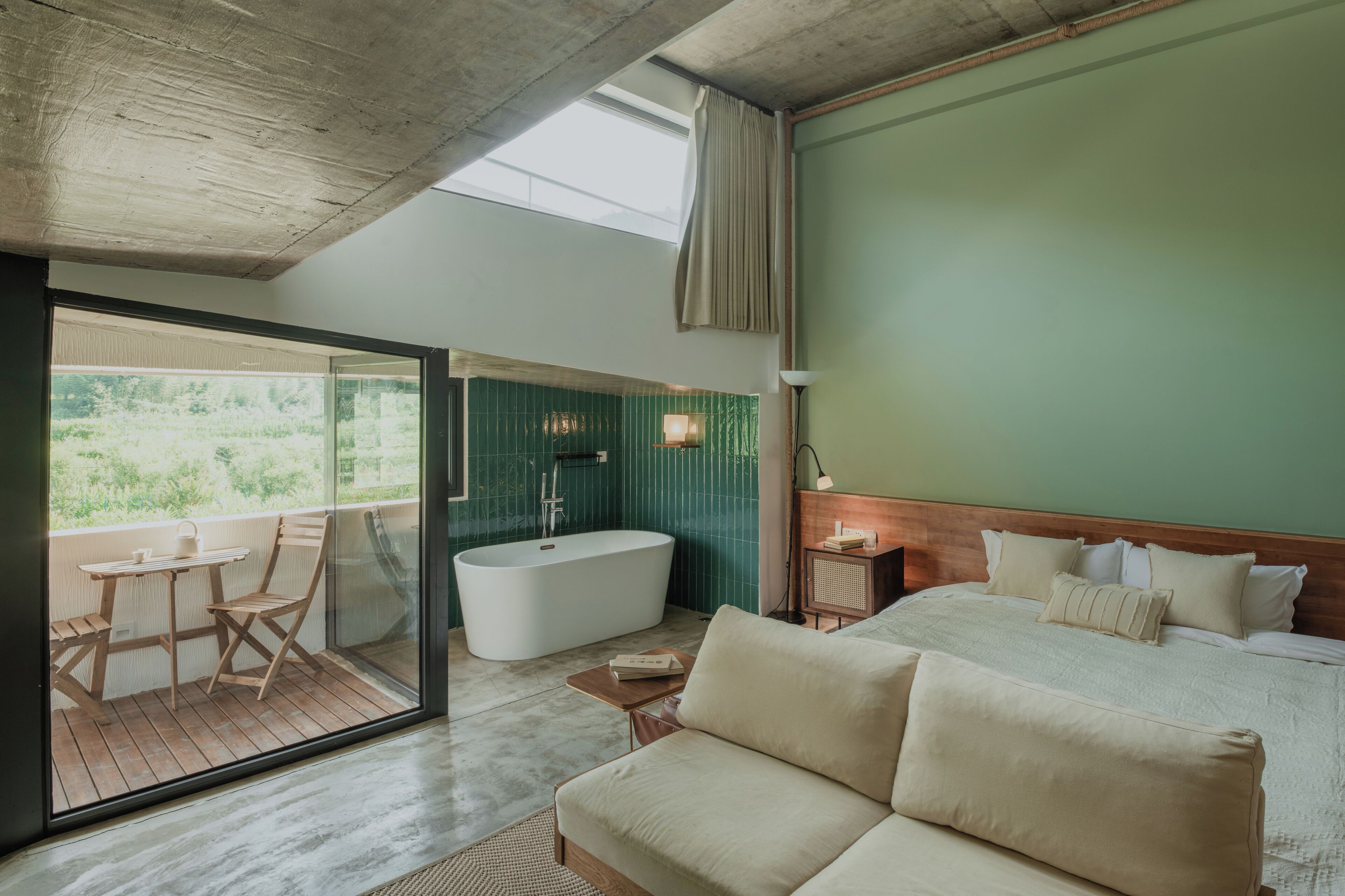 A modern bedroom with a large bed, a bathtub, a balcony with a table and chairs, and a view of greenery.
