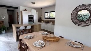 Un coin repas avec une longue table dressée pour quatre personnes, une cuisine en arrière-plan et un élément décoratif mural.