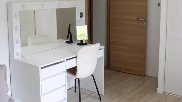 Une chambre avec un bureau blanc, une chaise et un lit. On y trouve une porte en bois et un mur blanc orné d’un miroir.