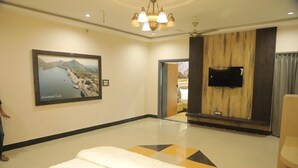 Quarto de hotel com uma cama, um quadro grande com uma paisagem de um lago, uma TV de tela plana presa a um painel de madeira e um lustre.