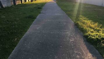 A concrete pathway flanked by grass and trees, leading towards a fence.