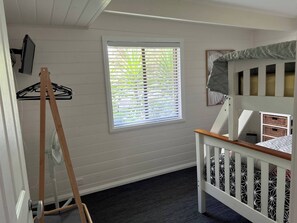 A room with a crib, a window with blinds, a fan, and a wall-mounted TV.