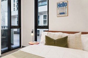 Ein Hotelzimmer mit einem Bett, einem Nachttisch, einer Lampe und einem großen Fenster mit Blick auf Gebäude.