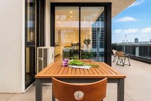 Une table en bois avec une chaise, une fenêtre donnant sur des immeubles et un balcon avec des chaises.