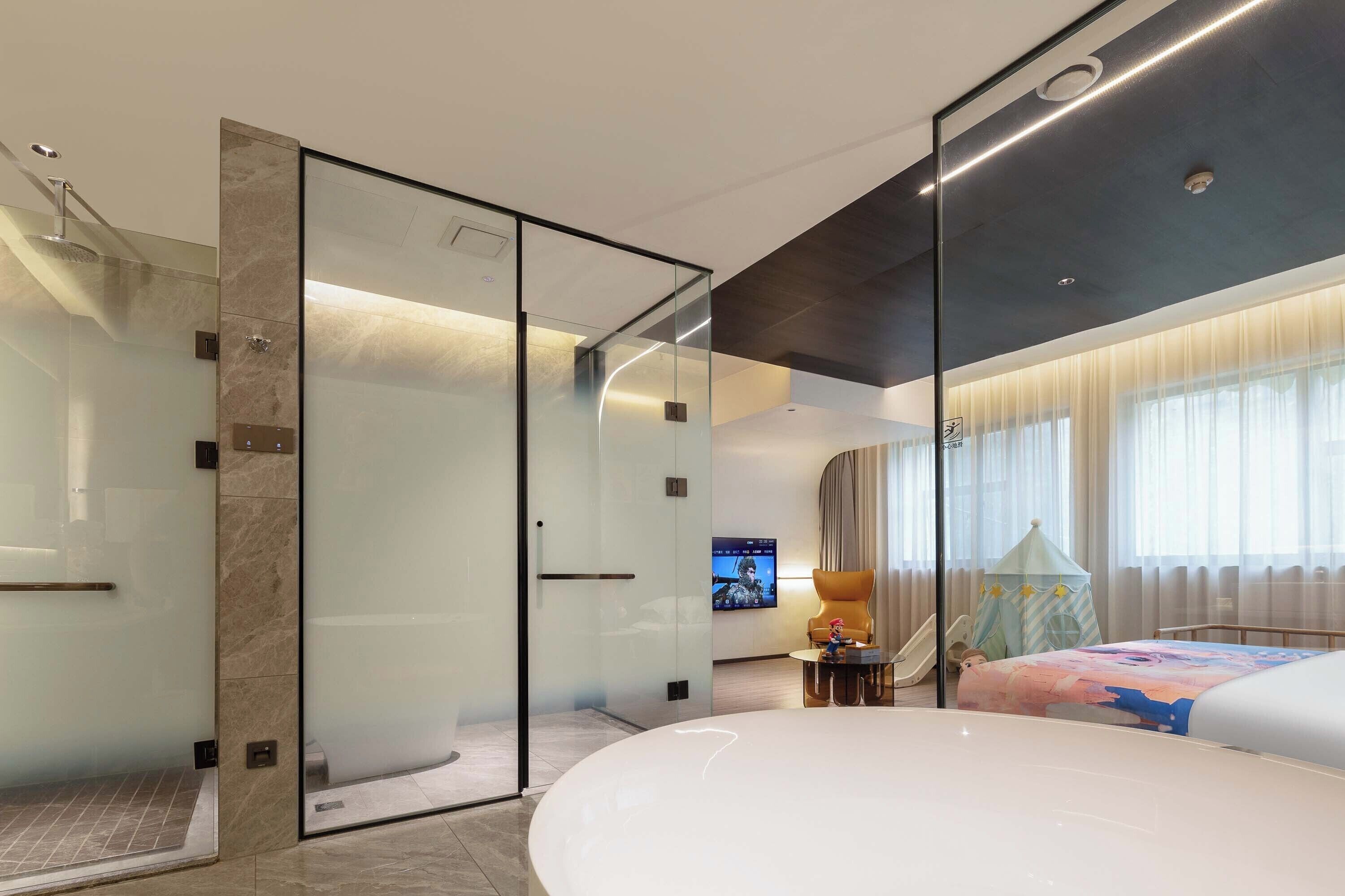 Una habitación de hotel moderna con ducha de vidrio, una bañera grande y un televisor montado en la pared.