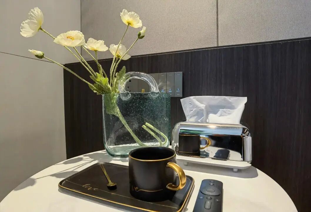 Una mesa con un jarrón de flores, una taza negra, una bandeja, un control remoto y una caja de pañuelos.