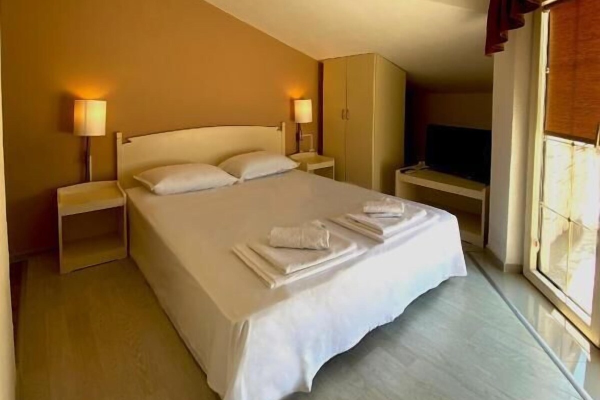 A hotel room with a large bed, bedside lamps, a TV, and a window with curtains.