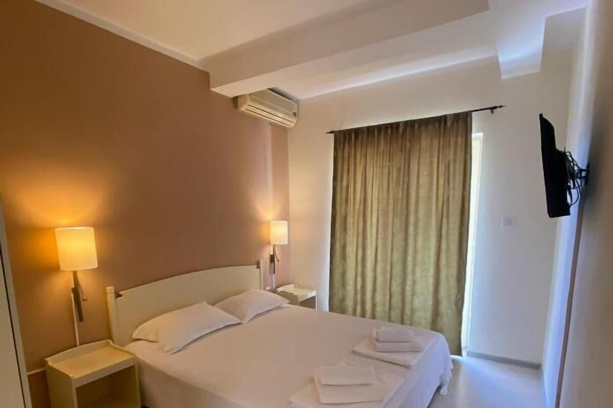 A hotel room with a bed, bedside lamps, a nightstand, a black box, and a window with curtains.