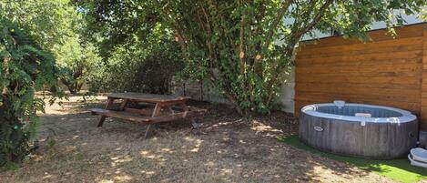 A wooden picnic table and a hot tub in a garden with trees and a wooden fence.