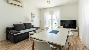 Ein modernes Wohnzimmer mit Sofa, Esstisch, Stühlen, einem Flachbildfernseher und einem großen Fenster mit Vorhängen.