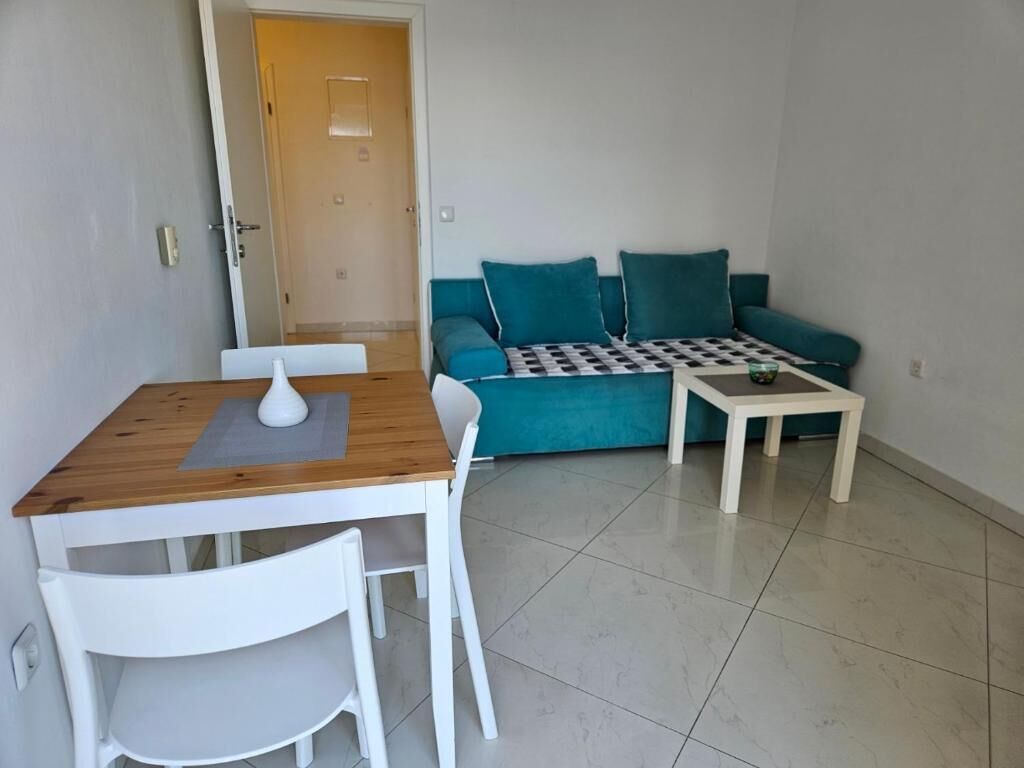 Un espacio de sala pequeño con una mesa de madera, sillas blancas, un sofá color turquesa y una pequeña mesa blanca.