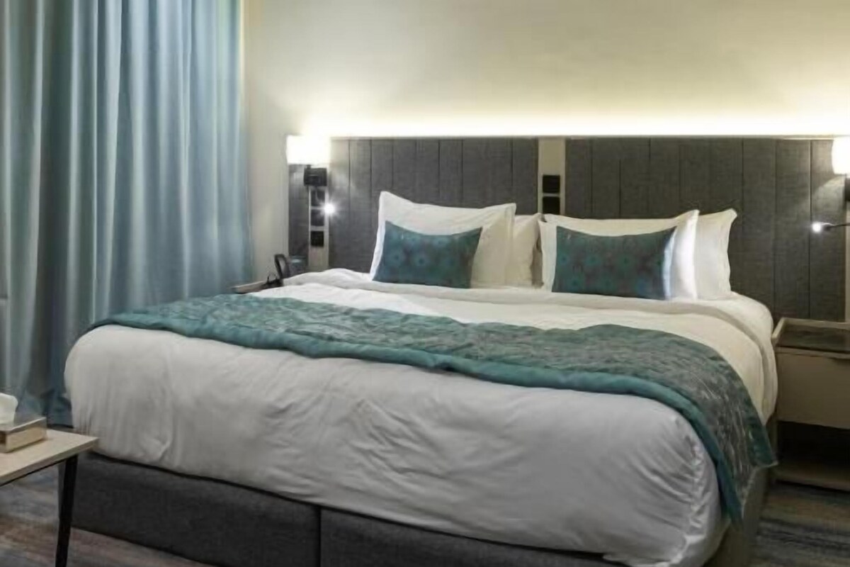 Quarto de hotel com uma cama grande, mesa de cabeceira e painel de cabeceira com iluminação embutida.