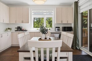 A modern kitchen with light wood cabinets, a dining table with white chairs, a coffee machine, and a view through a window. - LOB Wellness Retreat-Hot Tub-Sauna-Games-Fire Pit (Lake of Bays)