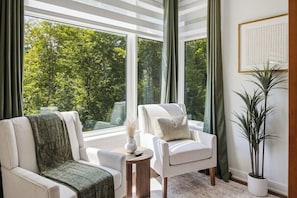 Ein Zimmer mit zwei weißen Sesseln, einem kleinen runden Tisch mit einer Vase und einer Fernbedienung, einem großen Fenster mit Blick auf Bäume und einer Zimmerpflanze.