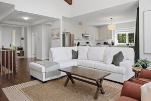 A modern living room with a white sectional sofa, a wooden coffee table, and a kitchen in the background. - LOB Wellness Retreat-Hot Tub-Sauna-Games-Fire Pit (Lake of Bays)