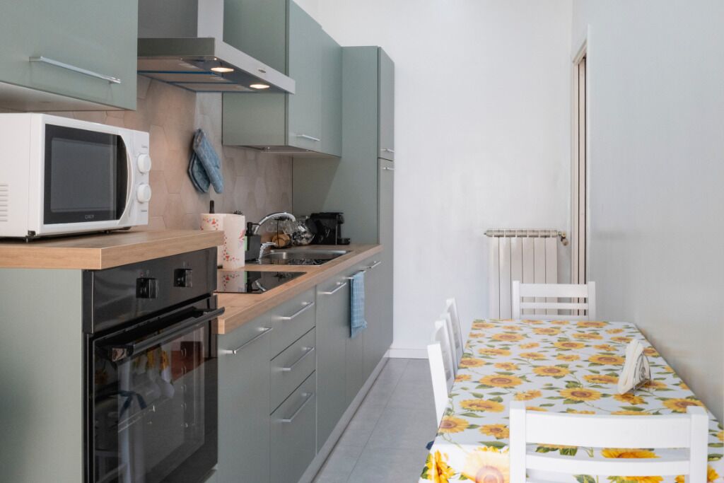 Een kleine keuken met een magnetron, oven en spoelbak. Er staat een eettafel met een tafelkleed met bloemenmotief en een stoel.