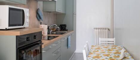 A compact kitchen with a microwave, oven, and sink. There's a dining table with a floral tablecloth and a chair.
