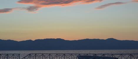 A sunset over a calm sea with a boat on the shore and mountains in the distance.